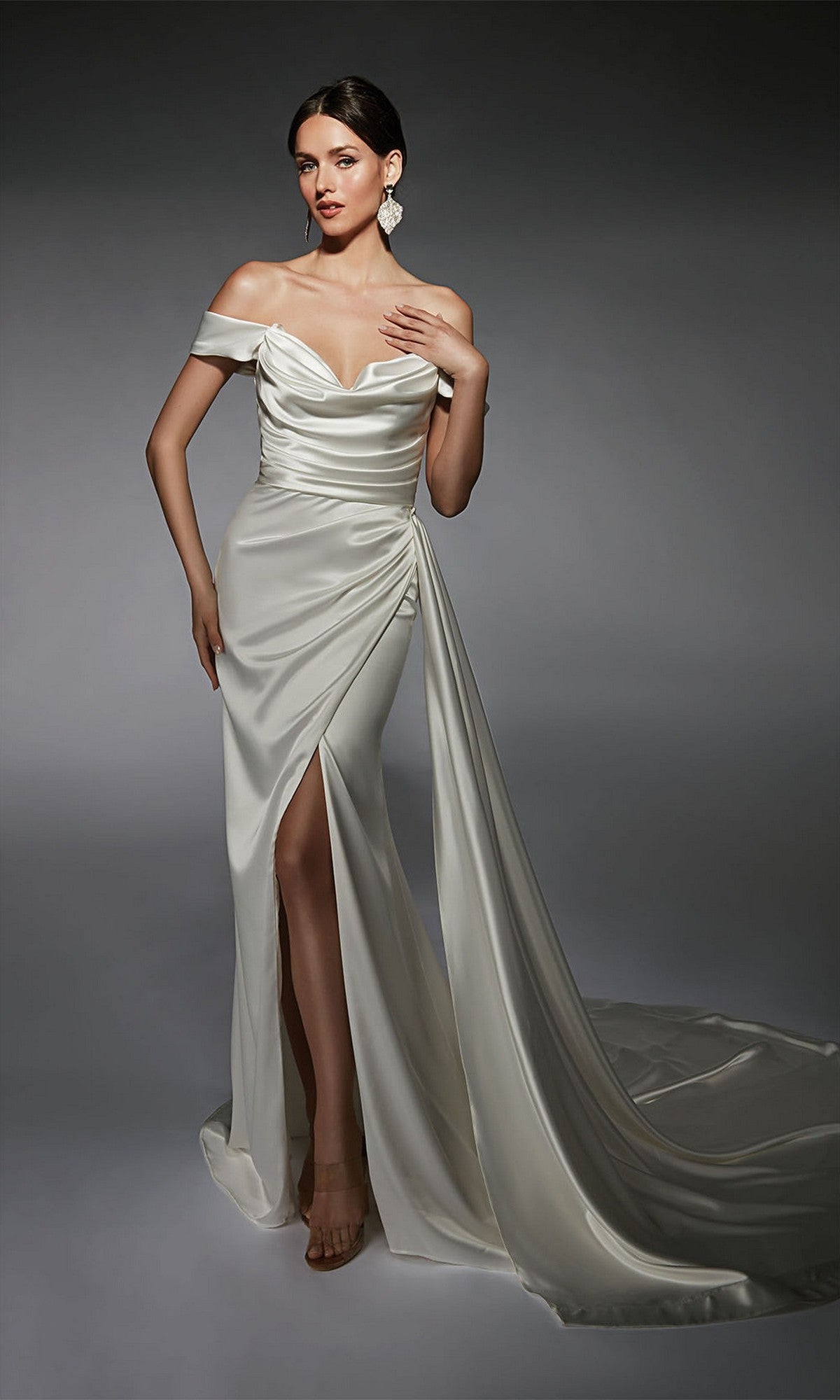 A woman stands against a gray background wearing the Long White Dress: Alyce 7106, featuring white satin fabric, a cowl neckline, off-the-shoulder sleeves, a thigh-high slit, and a long train.